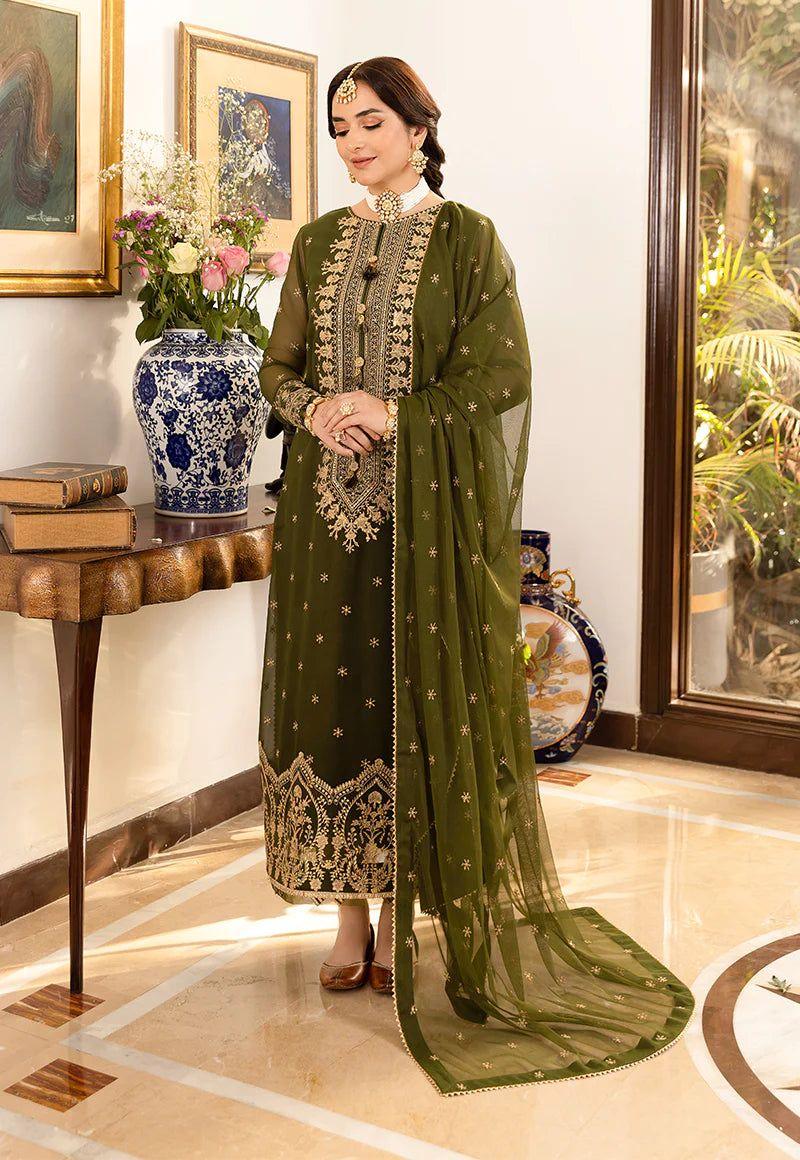 Woman in olive green embroidered traditional dress with matching dupatta, standing indoors near decorative vase and books