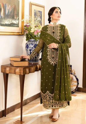 Woman in olive green embroidered traditional dress with matching dupatta, standing near ornate table with books and floral vase