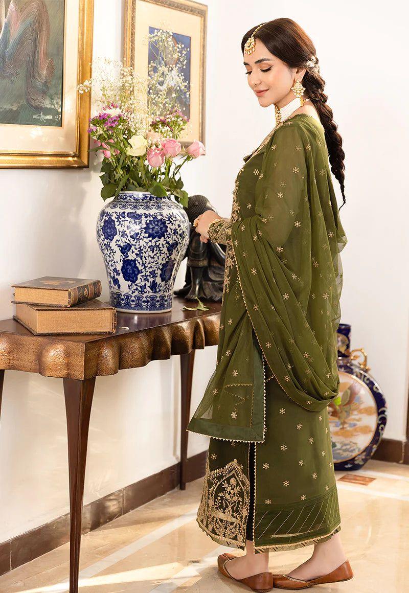 Woman in olive green embroidered traditional dress standing near table with blue floral vase and flowers
