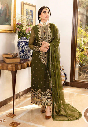 Woman in an olive green embroidered traditional dress with matching dupatta standing indoors near a wooden table with books and floral vase