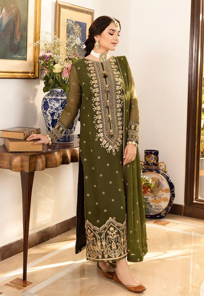 Woman wearing olive green embroidered traditional dress with gold jewelry standing indoors near wooden table and vase